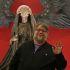 Guillermo del Toro sonríe y saluda frente a una figura monstruosa con alas negras y fondo rojo.