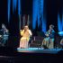 Vista general de Natalia Lafourcade sentada en el escenario cantando con micrófono, rodeada por su ensamble de cuerdas que incluye violines, violonchelo y contrabajo en el Teatro Metropólitan