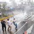 Jóvenes arrojan piedras hacia una línea de policías antimotines bajo chorros de agua a presión durante los disturbios en Guadalajara.