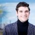 RJ Mitte, actor de Breaking Bad, sonríe con saco azul estampado y suéter negro de cuello alto frente a un fondo azul.