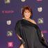 Alejandra Guzmán con cabello rojizo y vestido negro de un hombro, sonriendo ante un fondo morado de premios musicales.