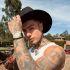 Naim Darrechi posa con sombrero negro, camiseta blanca y brazos tatuados en un espacio al aire libre con caballos.
