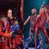 Ángela Aguilar canta con micrófono morado y ramo de flores, mientras Shakira baila con traje rojo durante un concierto.