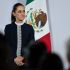 La presidenta Claudia Sheinbaum escucha atentamente durante su conferencia de prensa matutina, de pie frente a la bandera de México.
