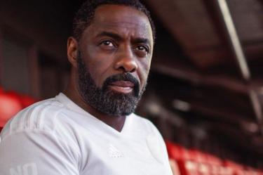 Idris Elba con playera blanca sentado en bancas rojas
