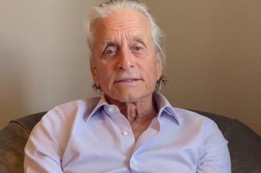 Michael Douglas con camisa morada, sentado en sillón