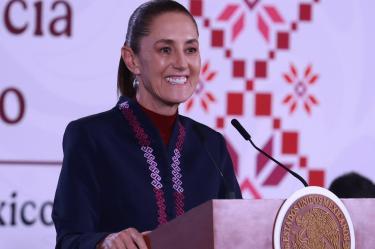 Fotografía en primer plano de Claudia Sheinbaum, Presidenta de México, sonriendo detrás de un atril oficial del gobierno con el escudo de los Estados Unidos Mexicanos y un fondo con patrones tradicionales rojos durante una conferencia de prensa.