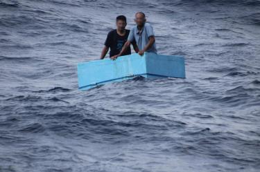 Dos hombres aferrados a una hielera rectangular de color azul claro, flotando a la deriva en el océano agitado.
