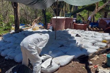 Perito con traje de protección inspecciona costales de precursores químicos bajo una lona en un mega laboratorio desmantelado.