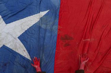 En esta imagen de archivo del 27 de octubre de 2019, una activista coloca las manos pintadas de rojo para simbolizar sangre sobre una bandera chilena, en homenaje a los manifestantes fallecidos en las protestas contra el gobierno, durante un concierto de protesta en Santiago de Chile. (AP Foto/Esteban Felix, Archivo)