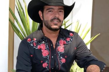 Pablo Montero de cuclillas con sombrero y camisa negra; mano sobre la pierna