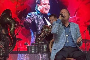 Julio Preciado con traje azul cantando, músicos e imagen de Juan Gabriel atrás