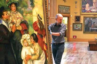Un curador de museo manipula cuidadosamente un gran cuadro de Pierre-Auguste Renoir en una galería de arte. La pintura muestra una escena familiar con varias figuras, incluyendo niños y adultos, en un entorno natural. Al fondo se aprecian más obras colgadas, lo que sugiere una exposición dedicada al impresionismo o al arte europeo del siglo XIX.