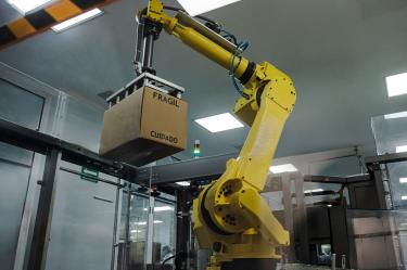 Brazo robótico industrial manipula una caja marcada como frágil dentro de una planta farmacéutica, representando la automatización de procesos, la innovación tecnológica y los estándares de seguridad en la producción de medicamentos.