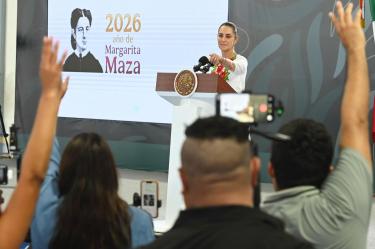Claudia Sheinbaum, Presidenta de México, preside una conferencia matutina en Tulum, Quintana Roo, con una pantalla al fondo que dice '2026, año de Margarita Maza'. Reporteros con las manos levantadas al frente
