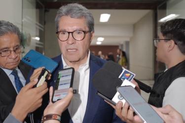 Ricardo Monreal responde preguntas de reporteros durante una entrevista en un pasillo, acompañado por otros asistentes y varios micrófonos.