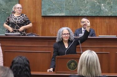 Primer plano de Graciela Domínguez Nava levantando la mano derecha mientras jura como gobernadora interina de Sinaloa en la tribuna del Congreso, con otros funcionarios de fondo.