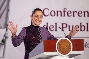 Fotografía de primer plano de la Presidenta de México, Claudia Sheinbaum Pardo, de pie detrás de un podio con el Escudo Nacional, sonriendo y gesticulando con las manos abiertas durante una conferencia de prensa matutina.