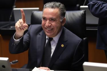 El senador Enrique Inzunza Cázarez sonríe y levanta el dedo índice en su escaño del Senado de la República.