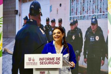 Clara Brugada habla ante un podio durante un informe de seguridad de la Ciudad de México, acompañada por imágenes de policías.