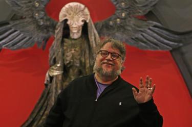 Guillermo del Toro sonríe y saluda frente a una figura monstruosa con alas negras y fondo rojo.