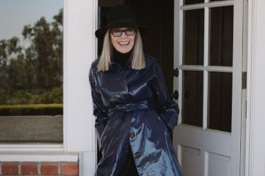 Diane Keaton con sombrero negro, lentes y abrigo azul brillante, sonriendo frente a una puerta blanca.