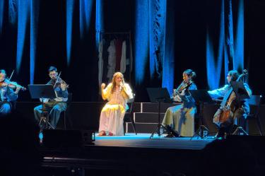Vista general de Natalia Lafourcade sentada en el escenario cantando con micrófono, rodeada por su ensamble de cuerdas que incluye violines, violonchelo y contrabajo en el Teatro Metropólitan