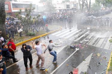 Jóvenes arrojan piedras hacia una línea de policías antimotines bajo chorros de agua a presión durante los disturbios en Guadalajara.
