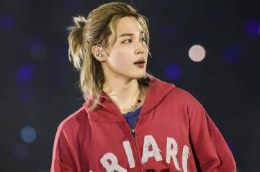 Jimin de BTS con sudadera roja, cabello rubio recogido y audífono de escenario durante un concierto.