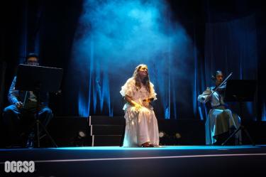 Natalia Lafourcade sentada en el escenario con un vestido blanco cantando sonriente, iluminada por un reflector azul, junto a dos violinistas.