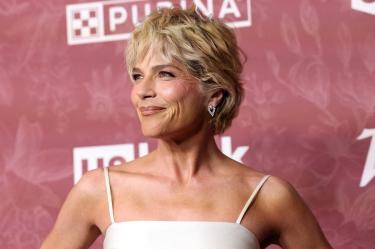 Selma Blair con cabello rubio corto, vestido blanco de tirantes y aretes plateados frente a un fondo rosa de Purina.