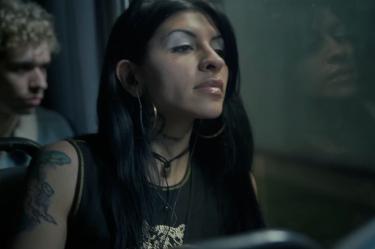 Cazzu con cabello negro, aretes de aro, audífonos y tatuajes visibles junto a una ventana en el videoclip de Con otra.