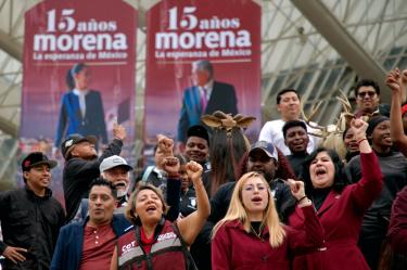Simpatizantes de Morena levantan los puños frente a lonas por el aniversario 15 del movimiento, con imágenes de Claudia Sheinbaum y Andrés Manuel López Obrador.