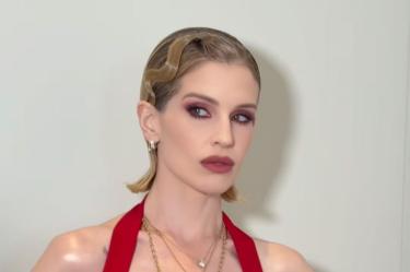 Kelly Osbourne posa con cabello rubio corto, vestido rojo, maquillaje rosa y collares dorados frente a un fondo claro.