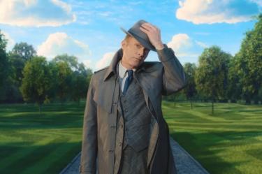 Paul Bettany como Vision en apariencia humana, con sombrero gris, traje y gabardina, en un parque de césped verde.