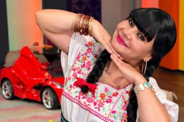 Bettina Salazar, Olga Sana, sonríe con blusa blanca bordada de flores, trenzas negras y las manos bajo el rostro.