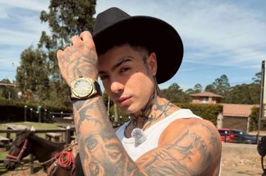 Naim Darrechi posa con sombrero negro, camiseta blanca y brazos tatuados en un espacio al aire libre con caballos.