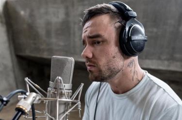 Liam Payne con audífonos negros y camiseta gris canta frente a un micrófono de estudio, con tatuajes visibles en el cuello.