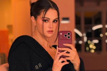 Selena Gomez se toma una selfie frente al espejo con un celular morado, bata negra y cabello recogido.