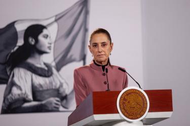 La Presidenta de México, Claudia Sheinbaum, vistiendo un saco rosa, habla desde un podio que porta el escudo de los Estados Unidos Mexicanos durante una conferencia matutina, con un mural con la bandera mexicana de fondo.
