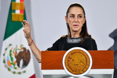 La presidenta Claudia Sheinbaum hablando desde el atril presidencial con el escudo de los Estados Unidos Mexicanos, teniendo la bandera de México de fondo.
