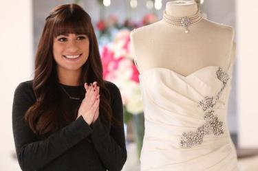Lea Michele como Rachel Berry en Glee, sonriente junto a un vestido de novia blanco con pedrería y collar de perlas.