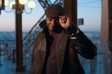 Omar Sy como Assane Diop en Lupin, con boina y abrigo oscuro, sonriendo mientras se acomoda la gorra frente a una ventana.