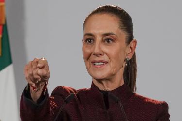 Claudia Sheinbaum hablando y señalando durante una conferencia de prensa con la bandera de México de fondo