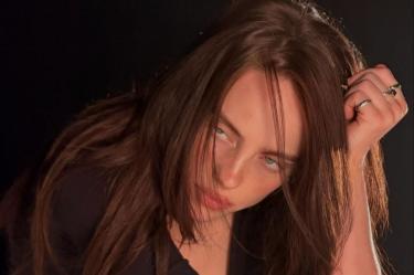 Billie Eilish posa con cabello largo castaño, ropa negra y una mano sobre la cabeza frente a un fondo oscuro.