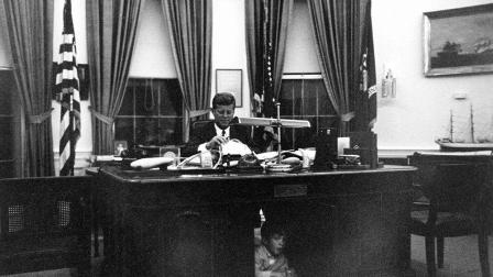 John F. Kennedy se sienta en su escritorio de la Oficina Oval, en la Casa Blanca, mientras su hijo Kennedy Jr. Mira por debajo.
