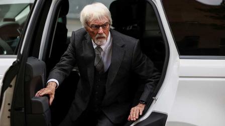 Bernie Ecclestone, vestido con un traje oscuro y corbata, descendiendo de un vehículo blanco. La imagen captura su mirada seria y su postura firme, destacando su papel como figura influyente en la historia de la Fórmula 1.