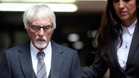Primer plano de Bernie Ecclestone, con traje oscuro, corbata gris y gafas, acompañado por una mujer vestida formalmente. Ambos caminan en un entorno profesional, destacando la seriedad y el carácter de la escena.