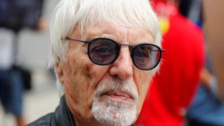 Retrato cercano de Bernie Ecclestone, con cabello blanco, barba bien recortada y gafas de sol oscuras, luciendo una chaqueta de cuero en un entorno lleno de actividad, característico de eventos de Fórmula 1.